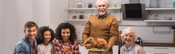 Encabezado del sitio web del hombre mayor sosteniendo pavo cerca de la familia multicultural en casa - foto de stock