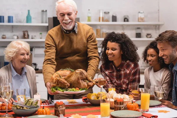 Focus selettivo della famiglia multietnica seduta vicino all'uomo anziano con tacchino in cucina — Stock Photo