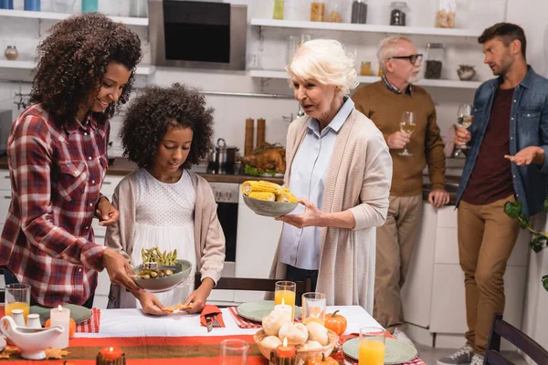 Enfoque selectivo de la anciana sosteniendo maíz cerca de la familia multiétnica y cena de acción de gracias en la mesa - foto de stock