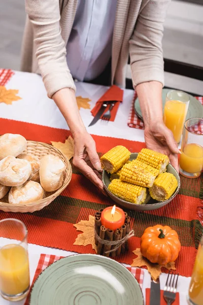 Cropped view of senior woman putting plate with corn on table near thanksgiving decorations — Stock Photo