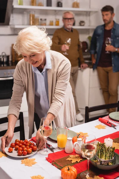 Enfoque selectivo de la mujer de pelo gris poniendo tomates cherry en la mesa durante el día de acción de gracias en casa - foto de stock