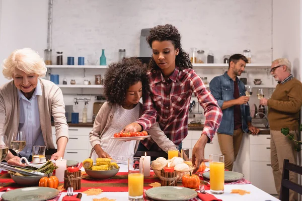 Foco seletivo de mulheres multiétnicas colocando comida na mesa perto da menina americana africana durante a ação de graças — Fotografia de Stock