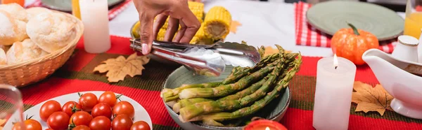Panoramic crop of african american woman holding tweezers near asparagus and candles on table during thanksgiving — Stock Photo