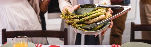 Horizontal image of elderly woman holding bowl of asparagus near multiethnic family during thanksgiving — Stock Photo