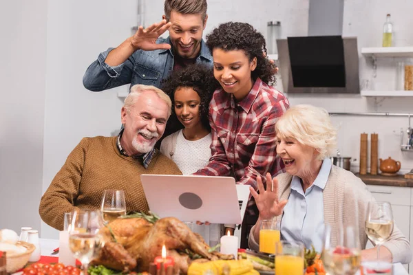 Enfoque selectivo de la familia multicultural que tiene videollamada en la computadora portátil cerca de la comida durante el Día de Acción de Gracias - foto de stock