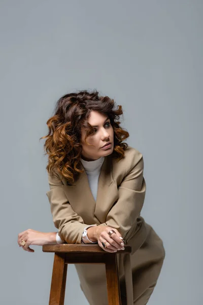 Elegant woman in beige suit posing on wooden chair and looking away isolated on grey — Stock Photo