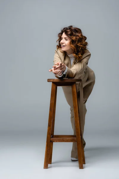 Элегантная женщина в бежевом костюме позирует на деревянном стуле и улыбается на сером — стоковое фото