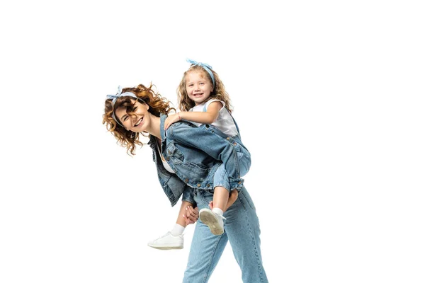 Side view of laughing mother piggybacking daughter in denim outfit isolated on white — Stock Photo
