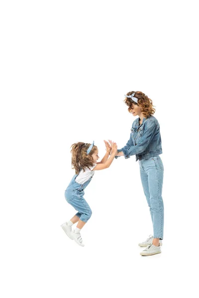 Vista laterale della figlia in abito denim saltando e battendo le mani con la madre isolata su bianco — Foto stock