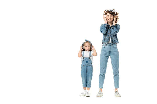 Madre e hija en trajes de mezclilla escuchando música en auriculares aislados en blanco - foto de stock