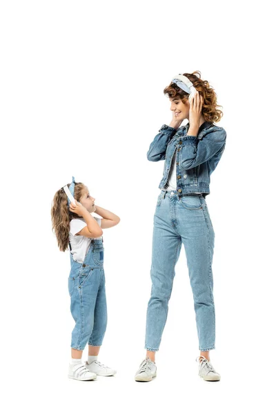 Mãe e filha em trajes de ganga ouvindo música em fones de ouvido e olhando uns para os outros isolados em branco — Fotografia de Stock