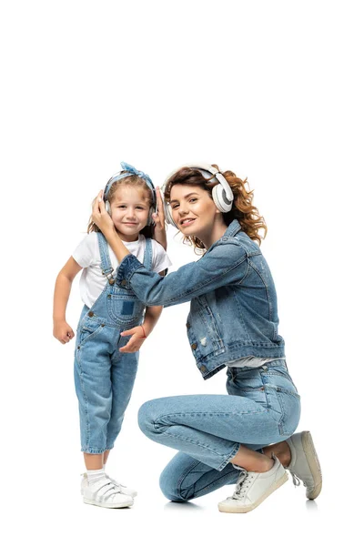 Madre e hija en trajes de mezclilla escuchando música en auriculares aislados en blanco - foto de stock