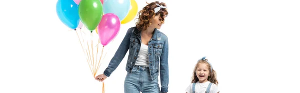 Madre e hija en trajes de mezclilla con globos de colores tomados de las manos aisladas en blanco, tiro panorámico - foto de stock