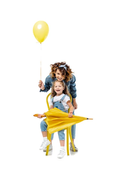 Mother with yellow balloon standing near daughter on chair with umbrella isolated on white — Stock Photo