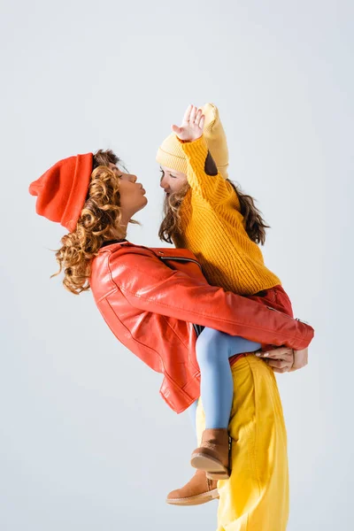 Vista lateral da mãe na roupa vermelha e amarela colorida segurando e beijando filha isolada no cinza — Fotografia de Stock