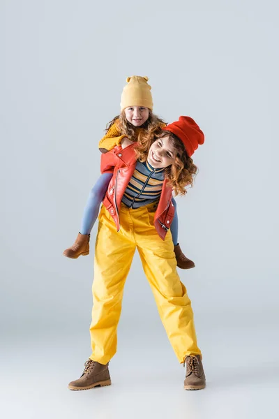 Mère et fille dans des tenues colorées rouges et jaunes piggyback sur fond gris — Photo de stock