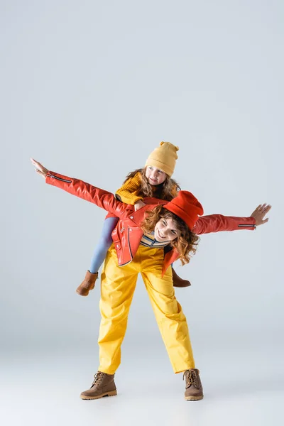 Mother in colorful red and yellow outfit imitating plane with daughter on back on grey background — Stock Photo