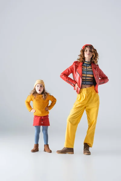 Madre e hija en trajes coloridos rojos y amarillos posando con las manos en las caderas sobre fondo gris - foto de stock