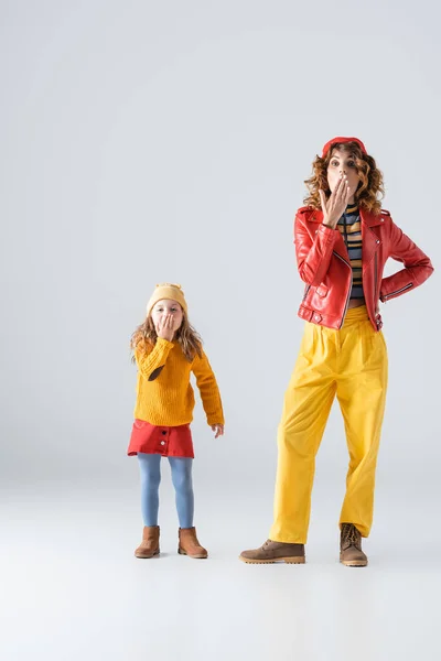 Impactados madre e hija en coloridos trajes rojos y amarillos cubriendo bocas sobre fondo gris - foto de stock