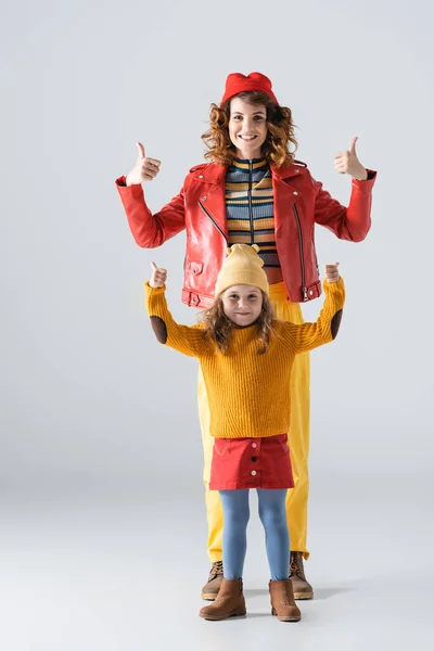 Madre e hija en trajes coloridos rojos y amarillos mostrando pulgares hacia arriba sobre fondo gris - foto de stock