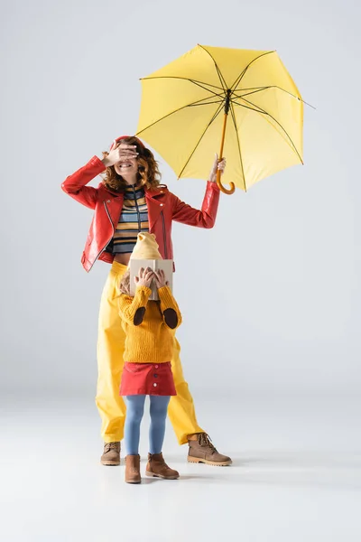 Madre e hija en trajes coloridos rojos y amarillos con paraguas y cubiertas de libros sobre fondo gris - foto de stock