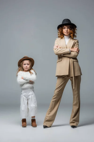 Elegante madre e hija en trajes blancos y beige posando con los brazos cruzados en gris - foto de stock