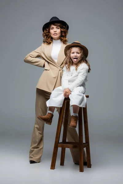 Mãe elegante posando perto da filha mostrando língua na cadeira no cinza — Fotografia de Stock