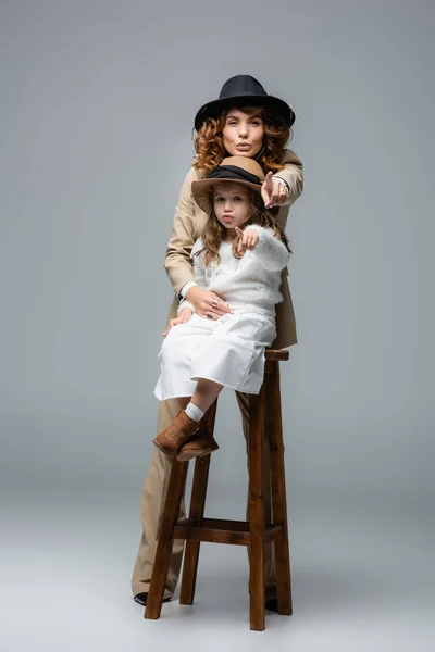 Елегантна мати позує біля дочки на стільці і вказує на камеру на сірий — стокове фото