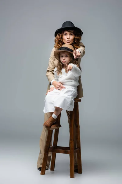 Elegant mother posing near daughter on chair and pointing at camera on grey — Stock Photo