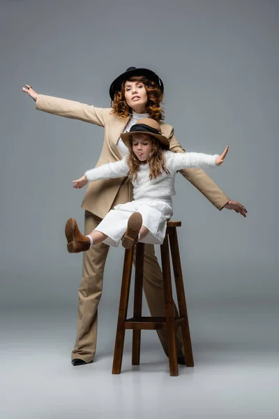 Elegant mother and daughter in white and beige outfits posing with outstretched hands on chair on grey — Stock Photo