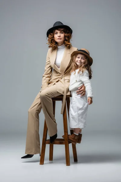 Elegante madre e figlia in abiti bianchi e beige in posa sulla sedia su grigio — Foto stock