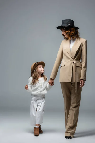Elegante madre e hija en trajes blancos y beige caminando y tomados de la mano en gris - foto de stock