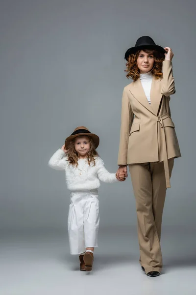 Elegante madre e hija en trajes blancos y beige y sombreros caminando y tomados de la mano en gris - foto de stock