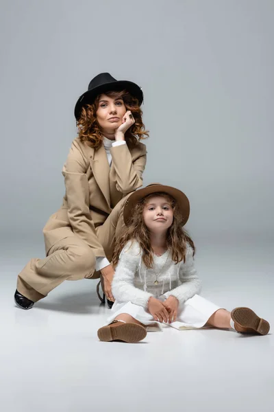 Elegante madre e hija en trajes y sombreros blancos y beige posando sobre gris - foto de stock