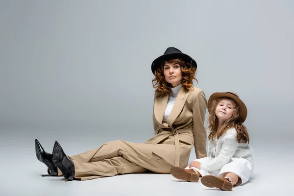 Elegant mother and daughter in white and beige outfits and hats posing on floor on grey background — Stock Photo
