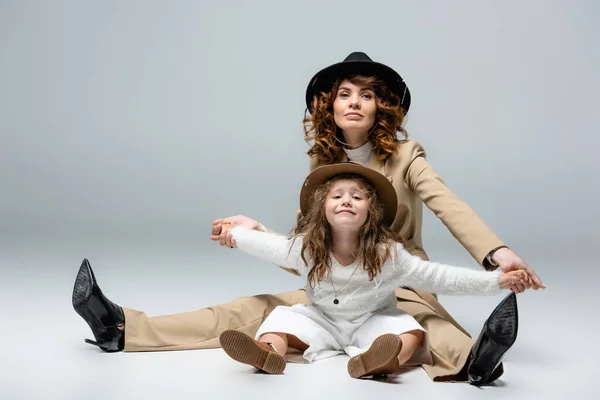 Elegante madre e hija en trajes blancos y beige y sombreros posando en el suelo sobre fondo gris - foto de stock