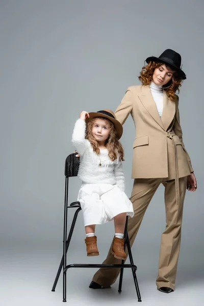 Elegante madre e hija en trajes blancos y beige y sombreros posando en silla sobre fondo gris - foto de stock