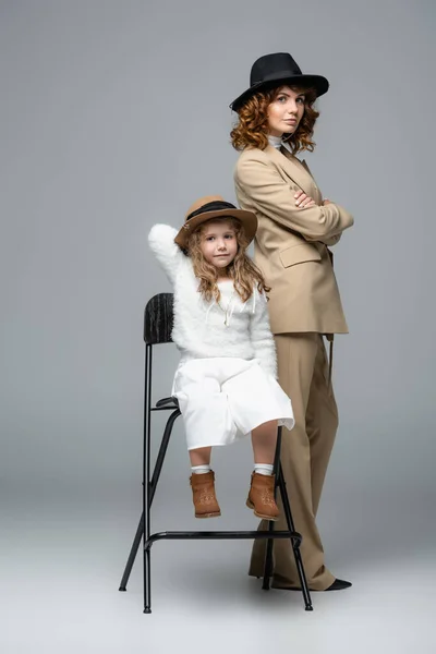 Elegante madre e hija en trajes blancos y beige y sombreros posando en silla sobre fondo gris - foto de stock