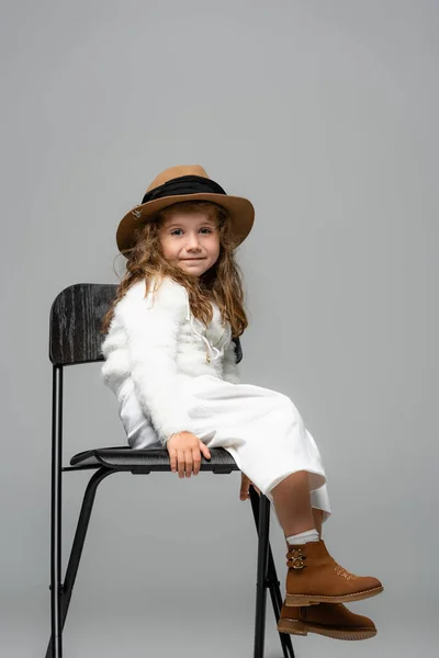 Elegante chica en traje blanco, sombrero marrón y botas posando en silla aislada en gris - foto de stock