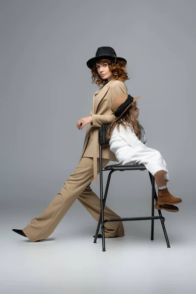 Side view of elegant mother and daughter in white and beige outfits and hats posing on chair on grey background — Stock Photo