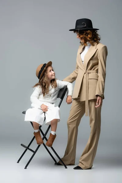 Elegant mother and daughter in white and beige outfits and hats posing on chair and looking at each other on grey background — Stock Photo