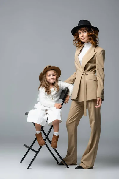 Elegant mother and daughter in white and beige outfits and hats posing on chair on grey background — Stock Photo