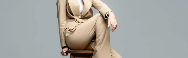 Vista recortada de mujer elegante en traje beige posando en silla de madera aislada en gris, tiro panorámico - foto de stock