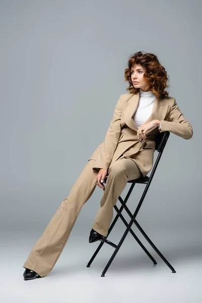 Elegante Frau im beigen Anzug posiert auf Stuhl vor grauem Hintergrund — Stockfoto