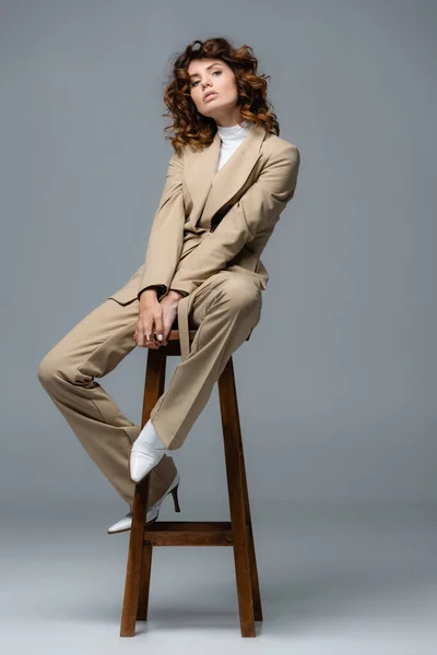 Elegante Frau im beigen Anzug posiert auf Holzstuhl vor grauem Hintergrund — Stockfoto