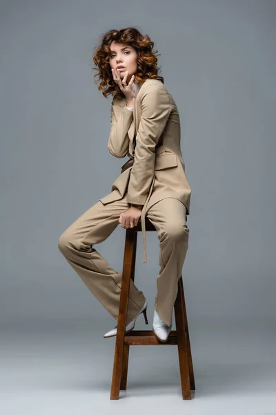 Элегантная женщина в бежевом костюме позирует на деревянном стуле на сером фоне — стоковое фото