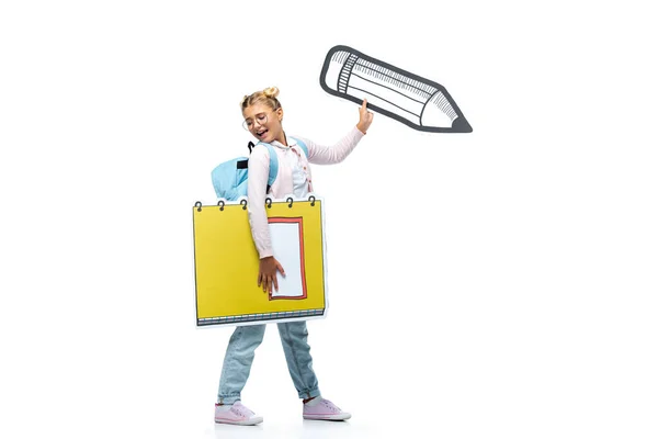 Full length view of excited schoolgirl in eyeglasses walking with paper cut copy book and pencil on white — Stock Photo