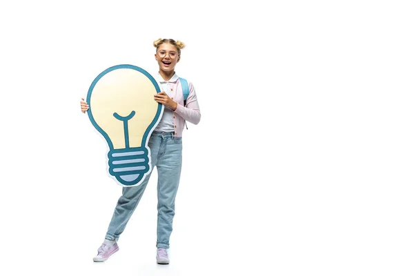 Full length view of excited schoolgirl in jeans holding paper cut light bulb and looking at camera on white — Stock Photo