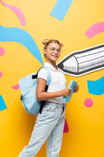 Joyful schoolchild with backpack looking at camera while holding book near multicolored elements and paper pencil on yellow — Stock Photo