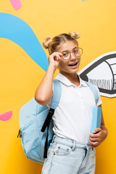 Aufgeregtes Schulkind, das Brillen anfasst und in die Kamera blickt, während es Buch in der Nähe bunter Elemente und Bleistift auf gelb hält — Stockfoto